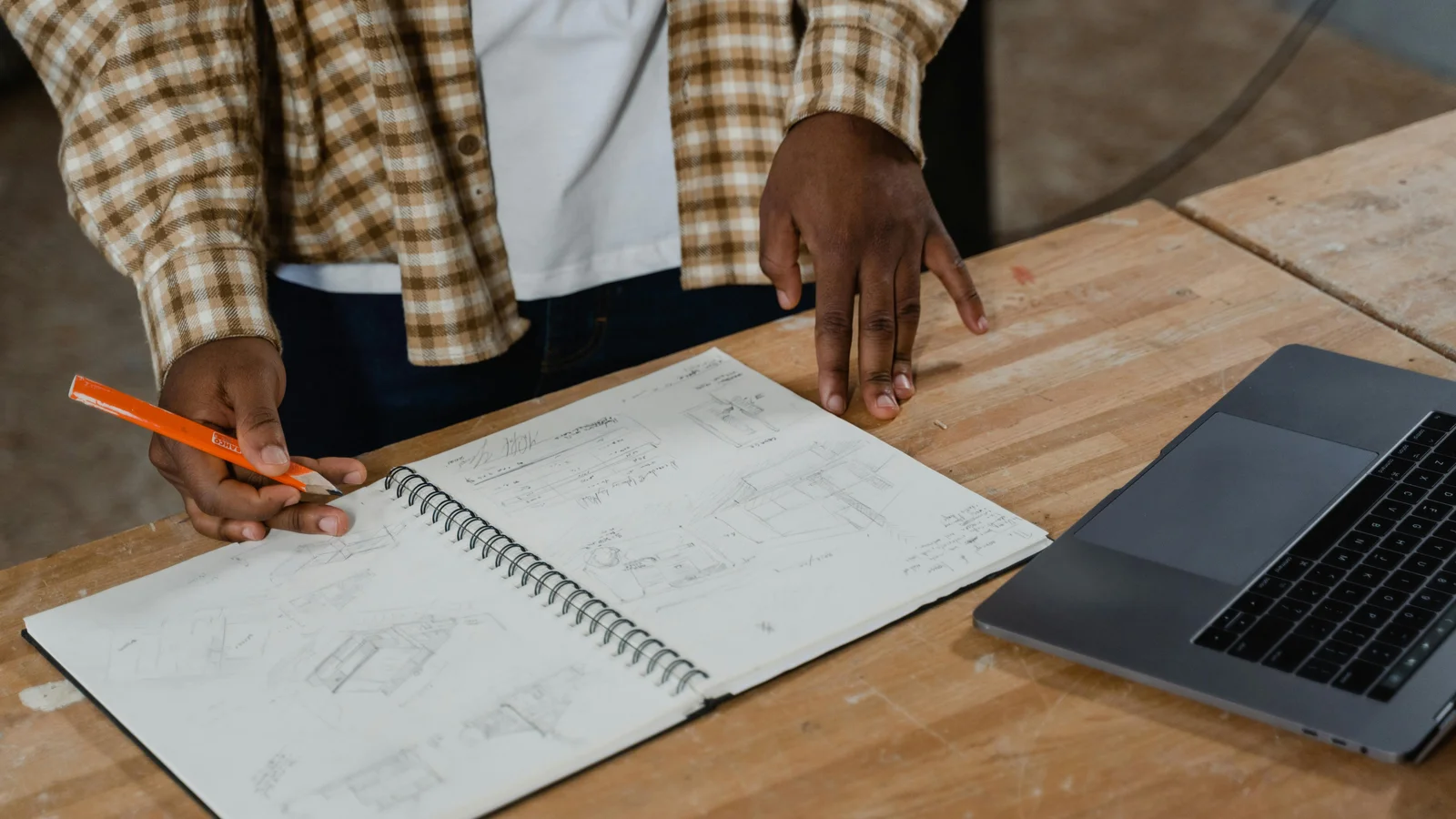 A craftsman at a workshop bench reviewing detailed product plans on paper next to an open laptop, deciding what the buyer needs to see before committing to a build.