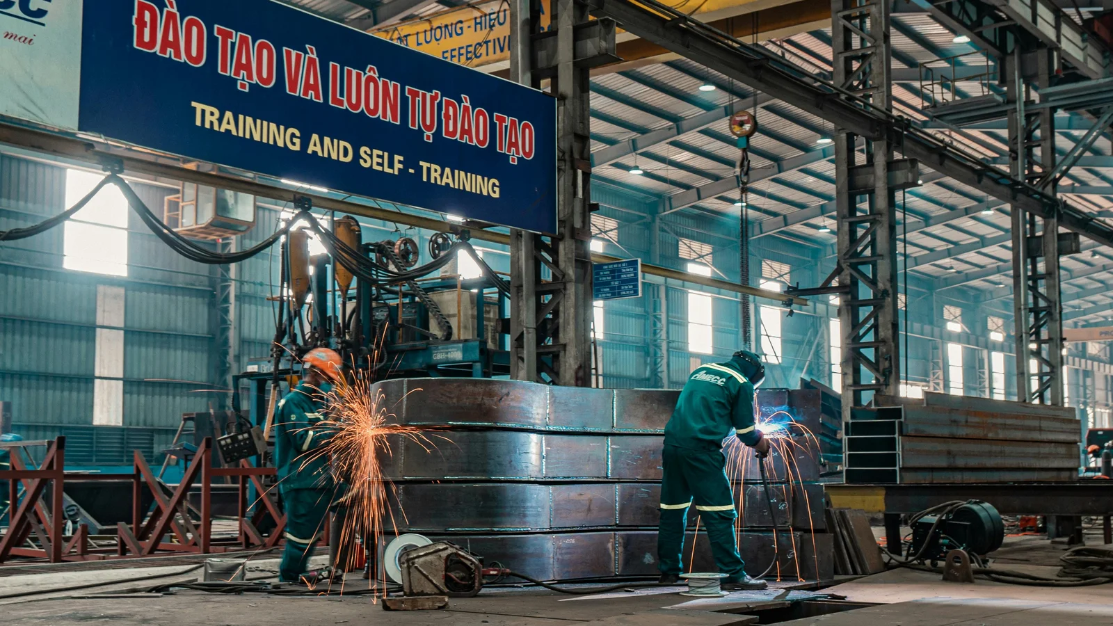 Two workers welding metal structures in a busy industrial fabrication shop, sparks flying, the kind of made-to-order manufacturing floor where every order has its own spec.