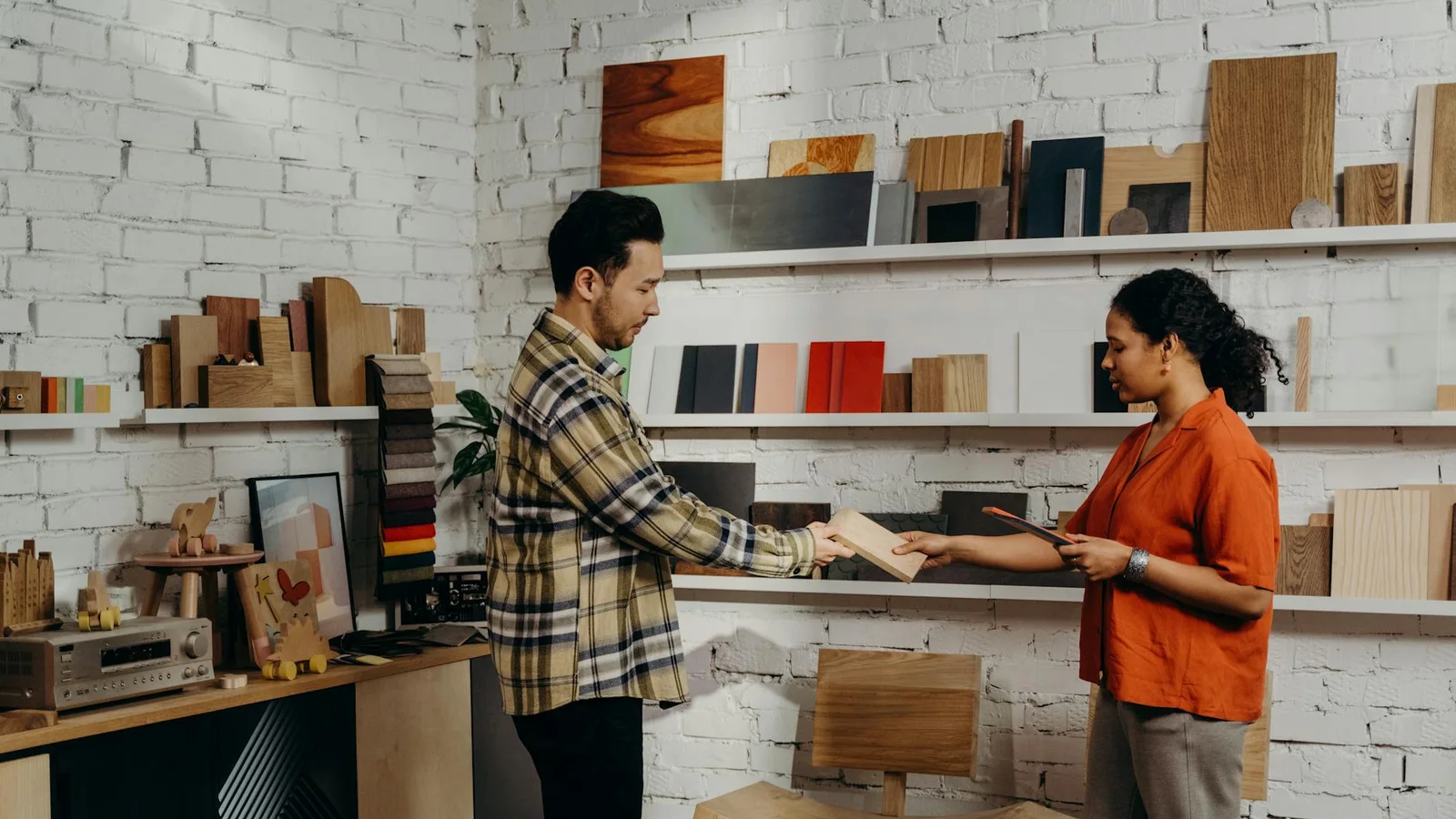 Two people in a materials showroom evaluating timber samples laid out on shelves, one holding a wood sample and the other a tablet.