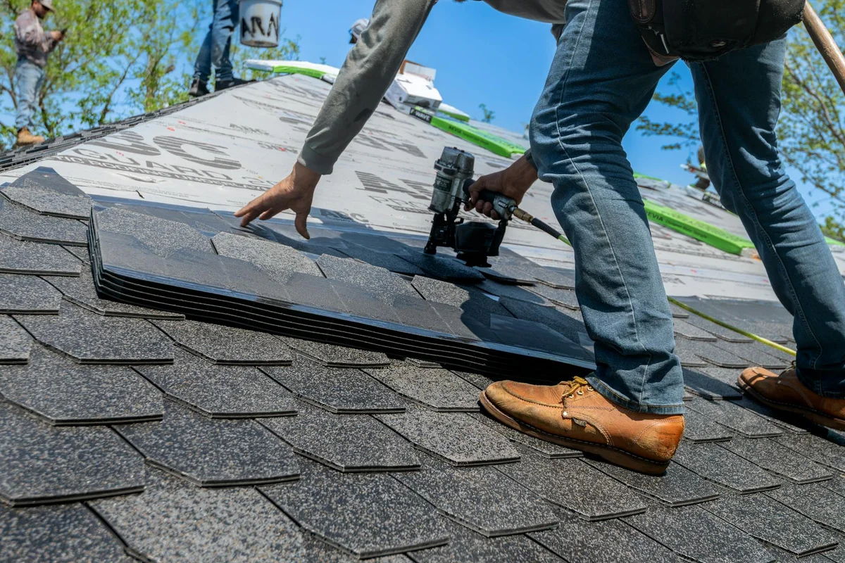 A roofer installing asphalt shingles on a residential roof with a nail gun, photographed from a low angle.