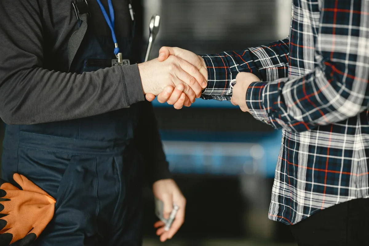 Close-up of a handshake between a contractor in a plaid shirt and a worker in a workshop uniform, tools visible at the side.