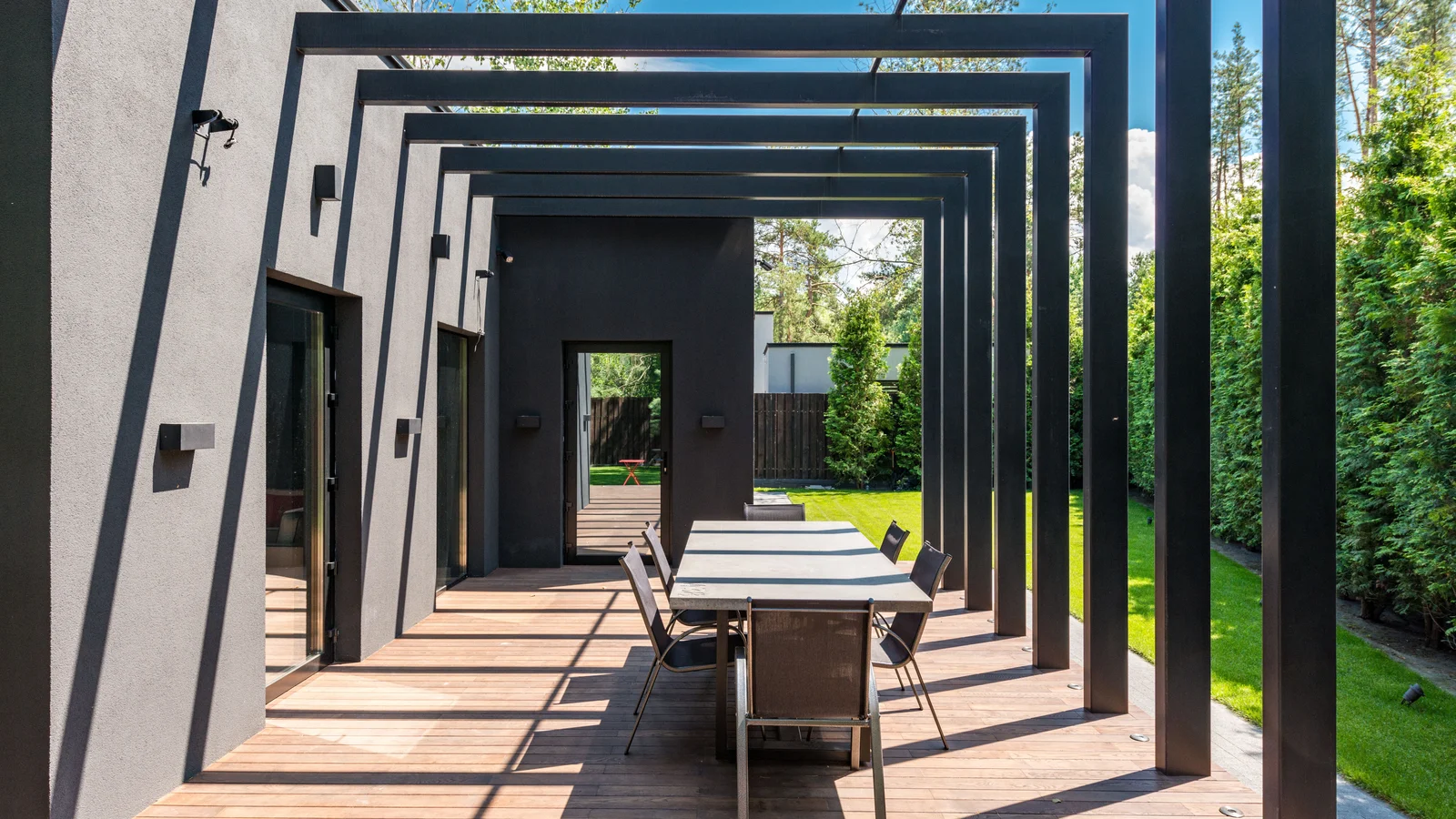 A modern black aluminium pergola with an open geometric frame over a timber deck and outdoor dining table, set against a dark contemporary house in clear daylight.