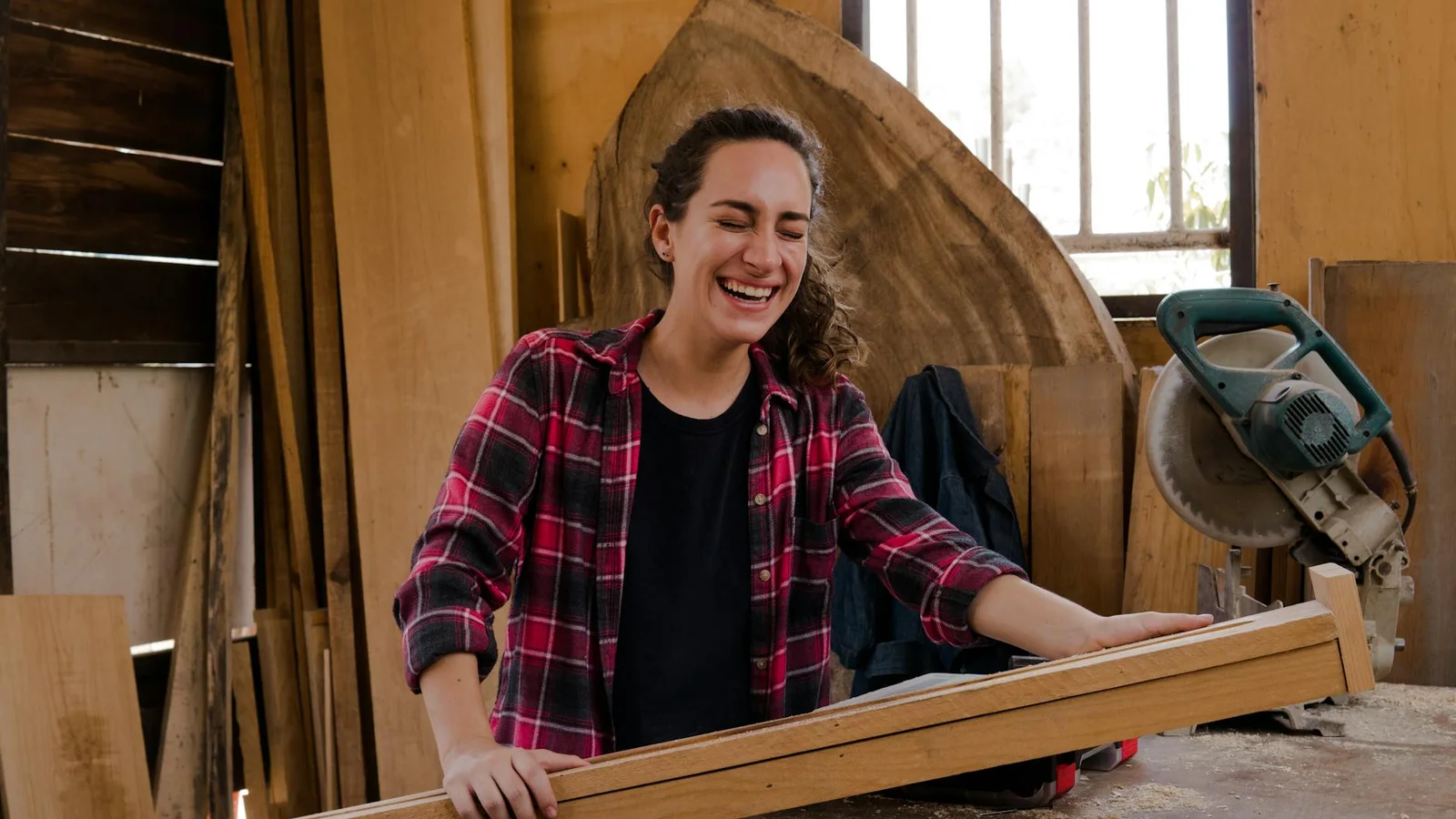A carpenter laughing in a wood workshop, holding a length of timber, with a mitre saw on the bench behind her.