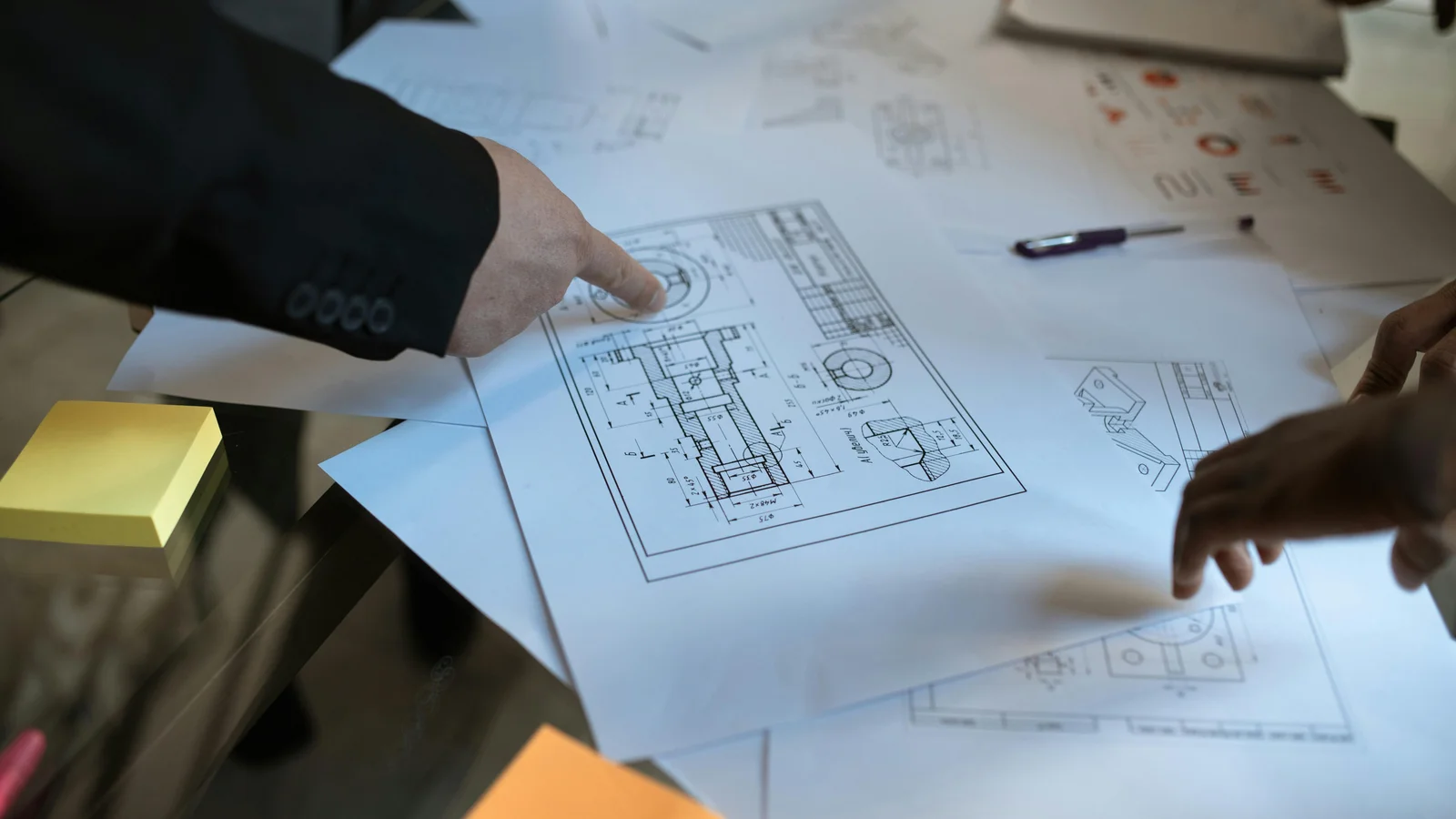 Two engineers leaning over a workspace, pointing at a printed blueprint as they walk through the configuration rules together.