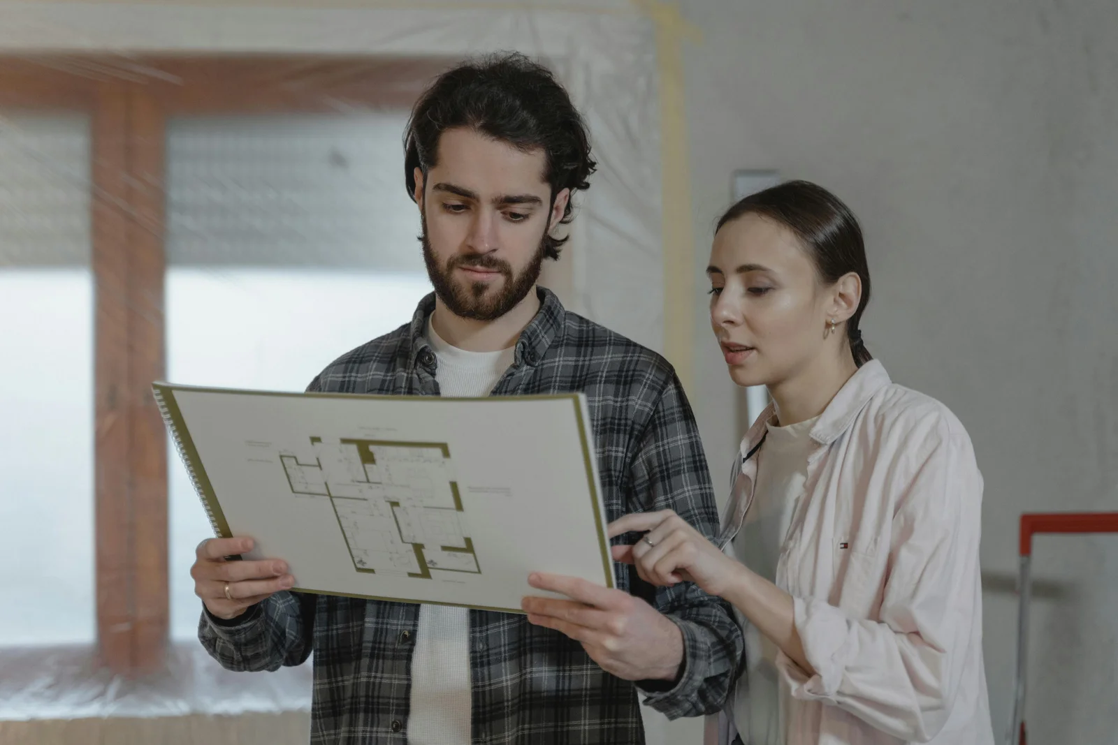 A homeowner couple working through a remodeling intake at the kitchen table, talking over a floor plan and a printed configuration before contacting a contractor.