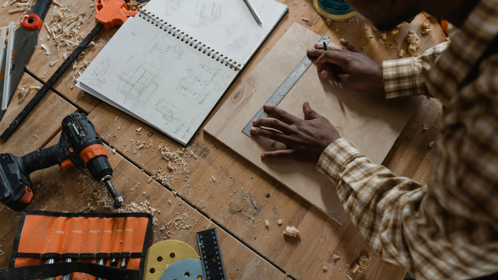 A pergola maker measuring a length of timber on a workshop bench beside a sketch and notebook, the desk where a hand-written quote takes shape.