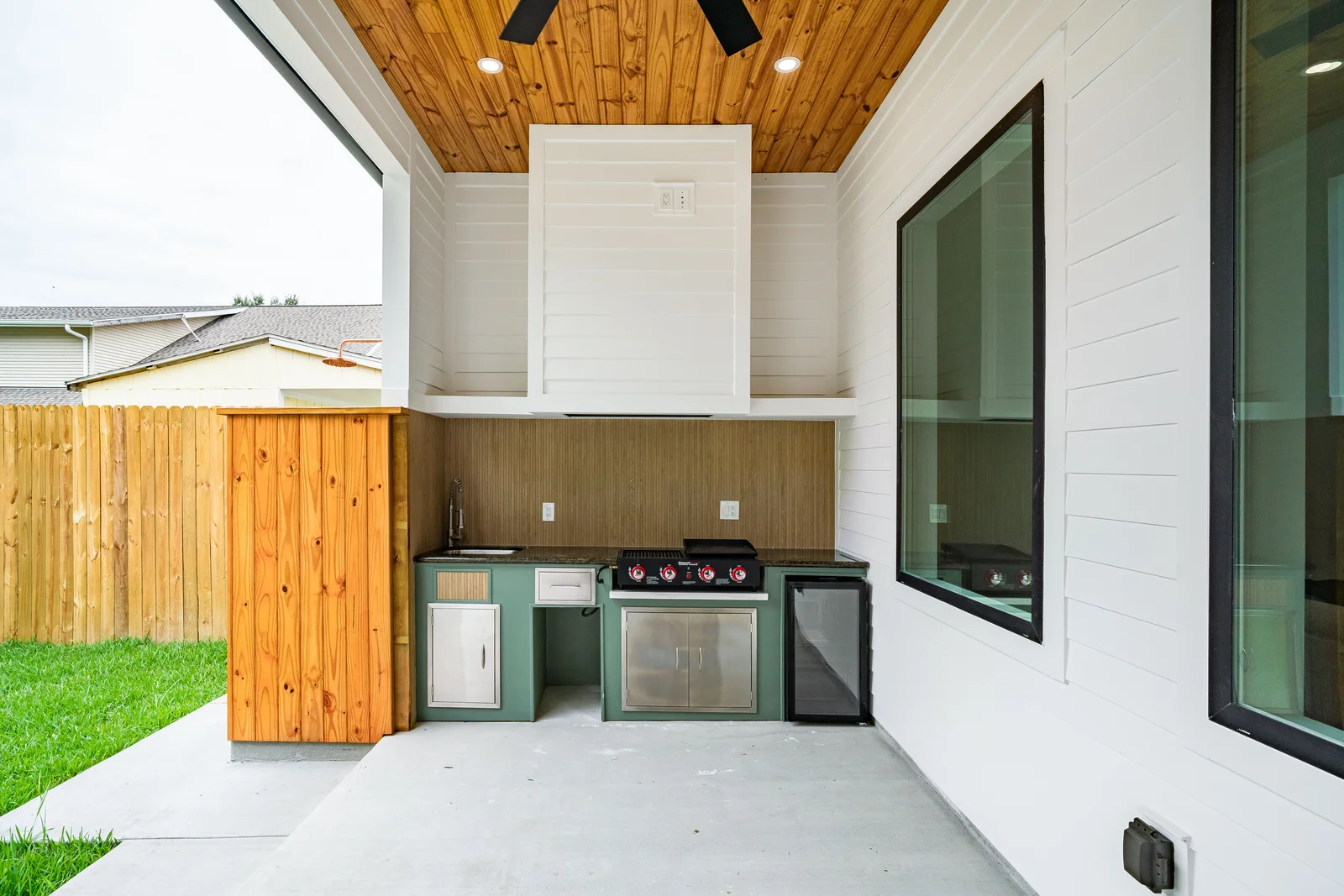 A built-in outdoor kitchen with a stainless grill, a fridge, and a stone counter under a timber-clad ceiling on a covered patio.