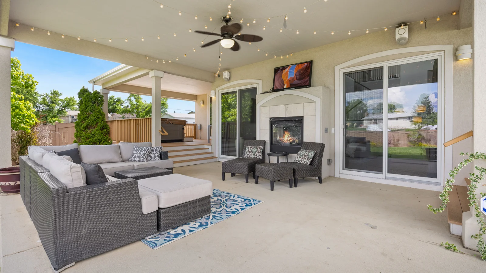 A covered outdoor living patio with a modern grey sofa set, a fireplace, and warm string lights at dusk in a residential backyard.