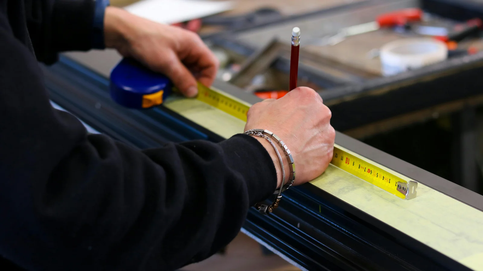 A workshop hand measuring an aluminium profile with a tape measure on a bench, a red pencil tucked behind the ear.