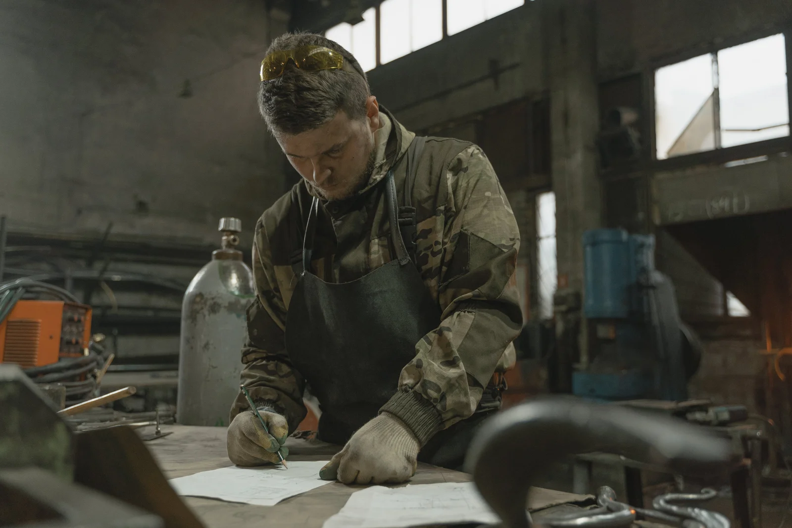 A man at a workbench writing on a notepad in a workshop, surrounded by metalwork tools, jotting down the spec for the next job.