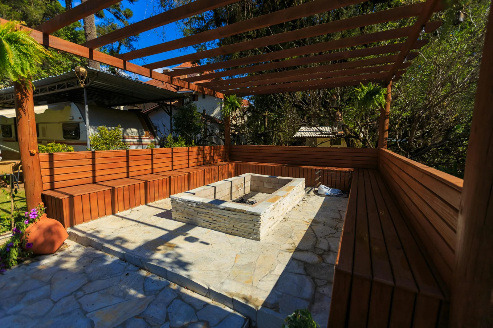 A finished outdoor patio with a stone fire pit and wooden benches set under a timber pergola, the build complete and ready for use.