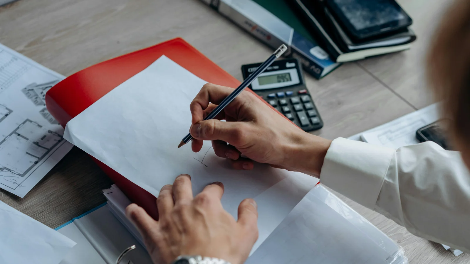 Close-up of hands writing on paper at a desk surrounded by a calculator, notebook, and rolled drawing plans, the scene of a quote being built by hand.