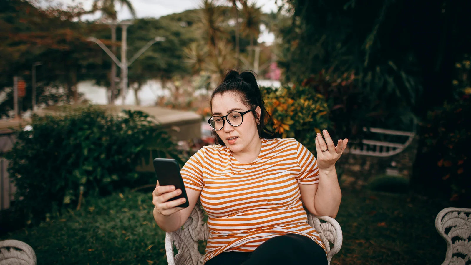 A woman sitting in a sunlit garden looking at her smartphone, the way an outdoor living buyer browses pergola options from the patio in the evening.