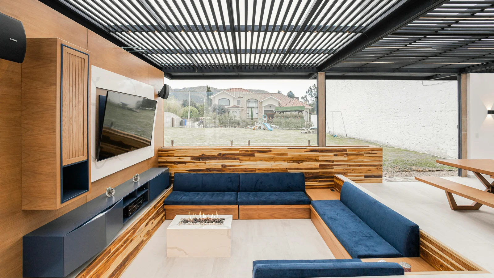 Anthracite aluminium louvered pergola roof seen from underneath, slats angled overhead above an outdoor living room with sofa, fireplace, and a wall-mounted television.