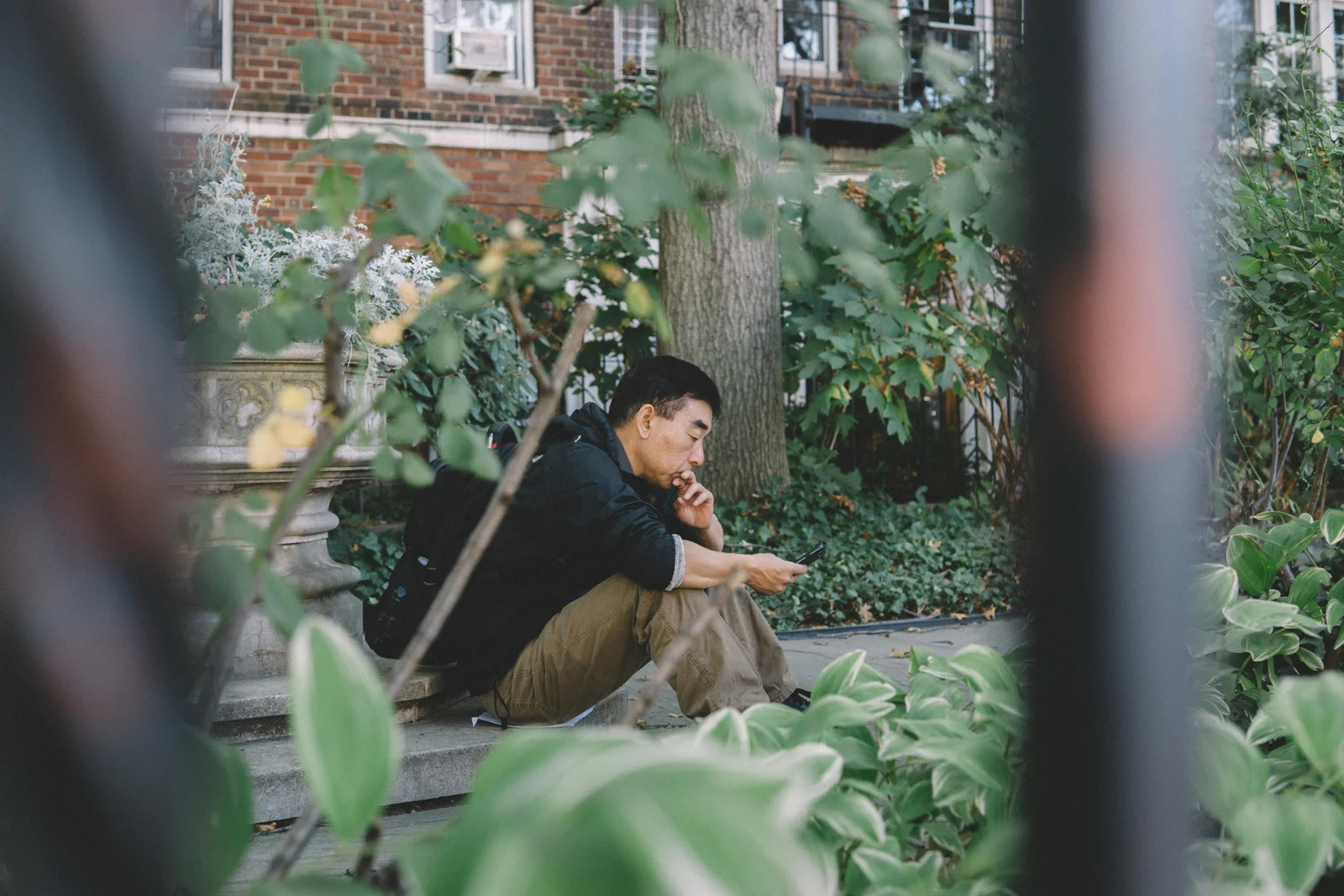 A young homeowner sitting outdoors among green plants, using a smartphone, the kind of moment when a residential buyer first opens a configurator on a manufacturer's website.