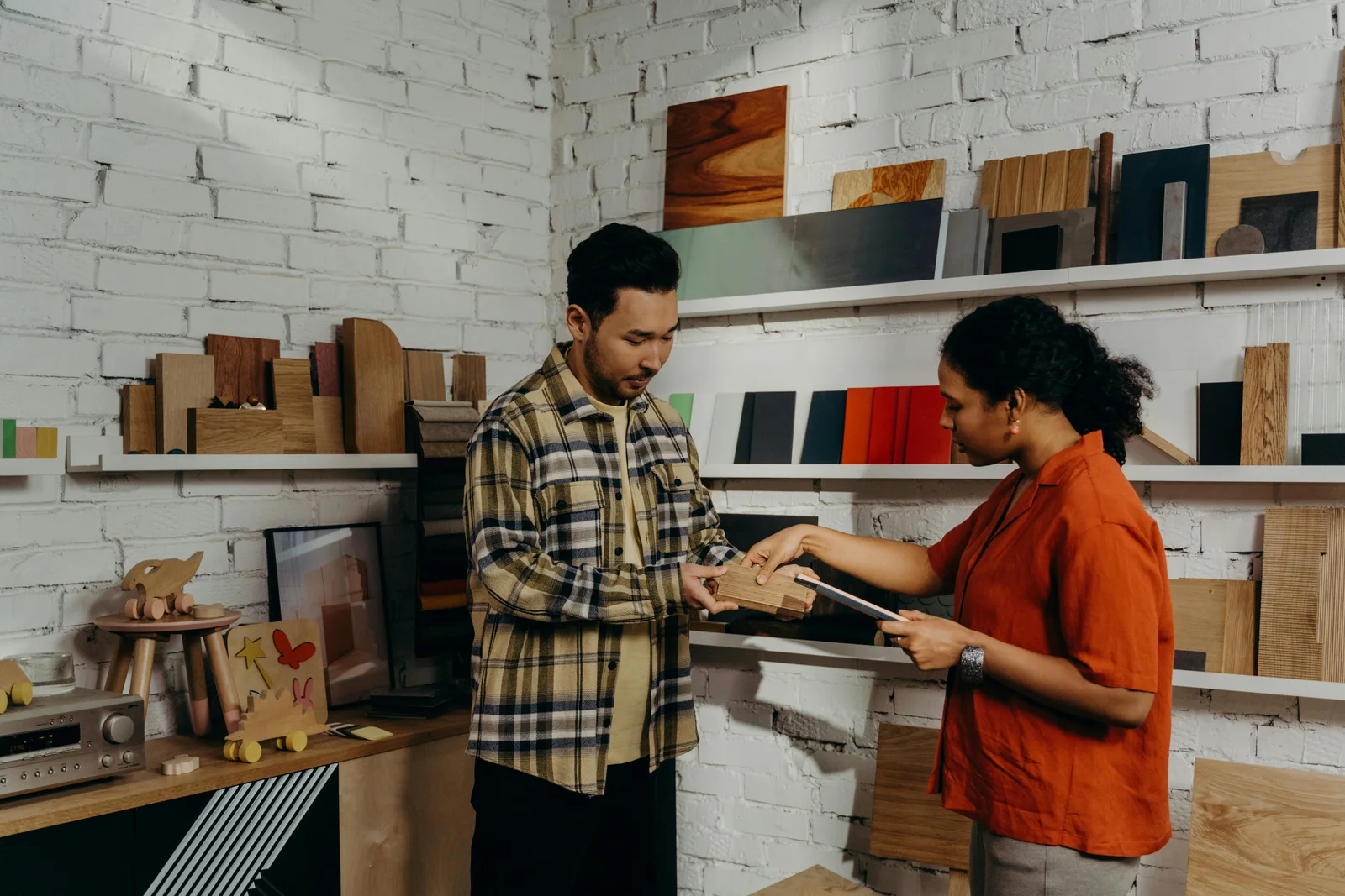 A man in a plaid shirt showing a wooden material sample to a woman in a contemporary furniture showroom setting.